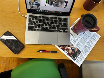 Brianna Adam's desk at Unparallel. pictured is a Mac book coffee cup phone pen and advertisement with local holiday events on it.